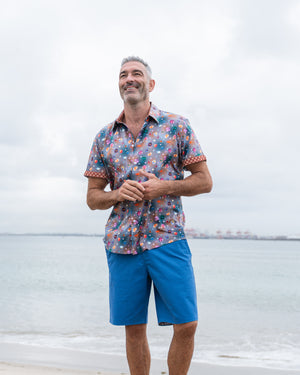 Man wearing a patterned shirt and blue chino shorts standing on a beach.