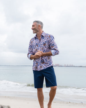 Man wearing a patterned shirt and navy shorts standing on a beach with ocean in the background