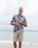 Man wearing a patterned shirt and beige shorts standing on a beach with a cloudy sky.