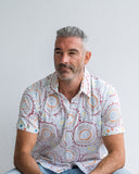 Man wearing a white patterned shirt sitting against a plain background