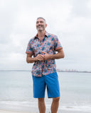 Man wearing a patterned shirt and blue chino shorts standing on a beach.