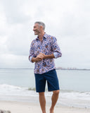 Man wearing a patterned shirt and navy shorts standing on a beach with ocean in the background