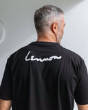 Man wearing a black t-shirt with 'Lennon' logo on the back against a white wall.